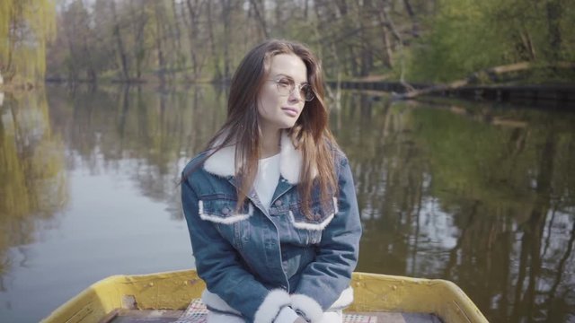 Portrait of a charming young woman in sunglasses and a denim jacket floating on a boat on a lake or river. Beautiful brunette is actively relaxing on a day off or traveling enjoying nature.