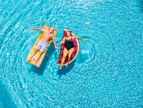 Above vertical view of people old senior couple taking hands with love and having fun on the blue clear swimming pool together enjoying the summer holiday vacation with trendy coloured lilos mattress