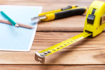Measuring tape tool with pencils, graph paper and stationery knife on wooden table.