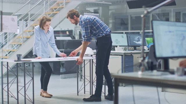 In the Industrial Engineering Facility: Young Female Designer Works with Chief Industrial Engineer, They have Discussion, Analyse and Correct Engine Design Technical Drafts that are Lying on the Table