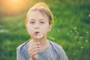 Happy child blowing dandelion flower outdoors. Girl having fun in spring park. Blurred green...