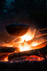 Food cooked on campfire in a large cooking pot, bright flames illuminating the dark scene and metal surface covered with soot, selective focus.