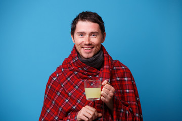 A wrapped in a blanket sick man is standing with cup of medicine in his hand.