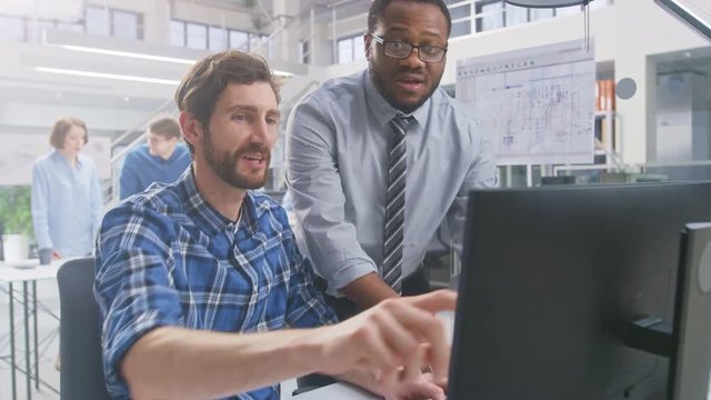 In the Industrial Engineering Facility: Portrait of the Handsome Male Engineer Working on Desktop Computer, Chief Engineer Stands Beside Overseeing Project. They Succeed and Cheerfully do High Five