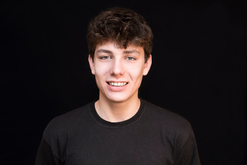 Fototapeta premium A teenager with dark hair in a dark shirt sincerely smiles on a dark background. The boy has a good mood