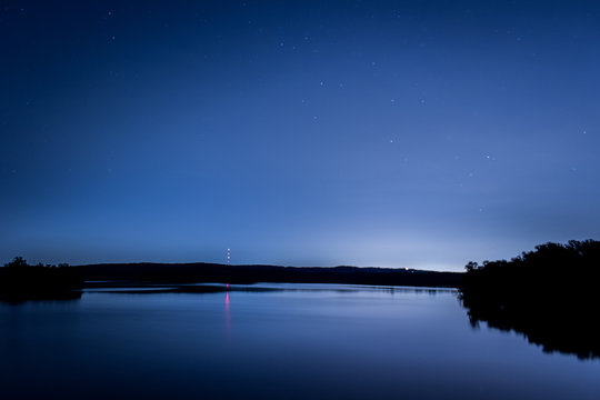 Star Trail Photo of Stars Above a Lake at Night - with Specks of Starlight in the Sky, Smooth Reflective Water, and Trees, Hills and a Red Beacon Light in the Background on a Clear Night in the Woods