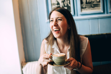 Stylish young woman laughs and drinks coffee in a cafe. Scarred with foam