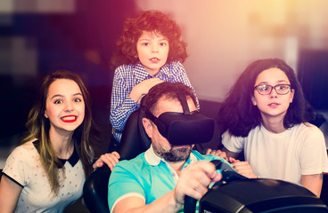 technology, gaming, entertainment and people concept. Happy young man is playing racing videogame in 3D virtual reality simulator using headset. Happy family having fun together.