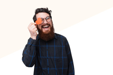 Photo of happy bearded guy iscovering eye with little paper heart, smiling over white background