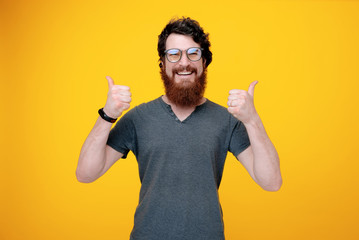 Happy handsome man with beard, wearing glasses, and showing thumbs up  over isolated yellow wall