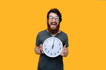 Excitd bearded guy with galsses, holding a clock, standing over yellow background