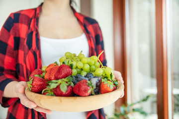 Woman holds wooden bowl with green grapes, strawberries and blueberries with window in the background.