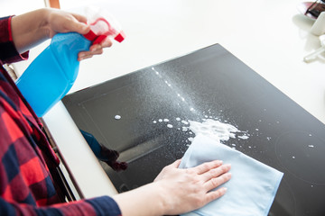 Woman splashes cleaning foam from blue bottle on the induction hob and wipes it using blue cloth.