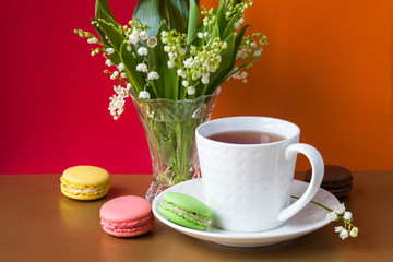 French multicolored cakes macarons and a cup of tea. A bouquet of lily of the valley in a crystal vase.
