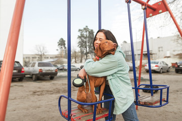 Naklejka premium Happy girl hugs a beautiful young dog sitting on a swing. Portrait of a happy owner with your favorite dog. Woman rests with the dog in the weekend