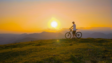Fototapeta na wymiar SUN FLARE: Cross country biker stands on top of a hill and watches the sunset.