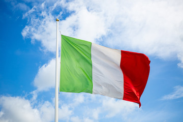 Waving Italian flag on a flagpole with an intense blue sky and clouds in the background slightly defocused