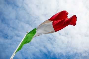 Waving Italian flag on a flagpole seen from below with a blue sky and clouds in the background slightly defocused