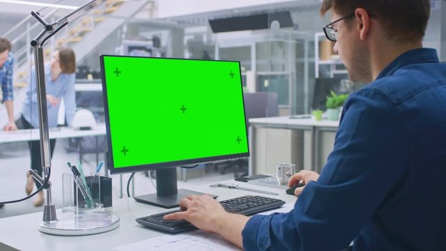 Over the Shoulder Shot of Engineer Working with Green Mock-up Screen Desktop Computer. In the Background Engineering Facility with Specialists Working on Blueprints and Drawings with Industrial Design
