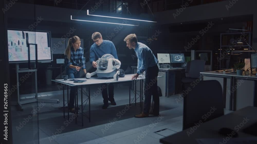 Leinwandbilder Late at Night in Robotics Engineering Facility Three Technical Engineers Talk and Work on a Wheeled Robot Prototype. In the Background High Tech Research Center with Screens Showing Industrial Design