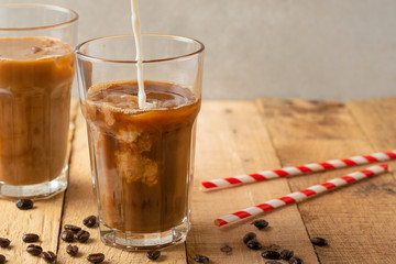 Cold coffee pouring in cream, milk in transparent glasses with ice and straws, on a wooden background, a cooling drink, refreshing