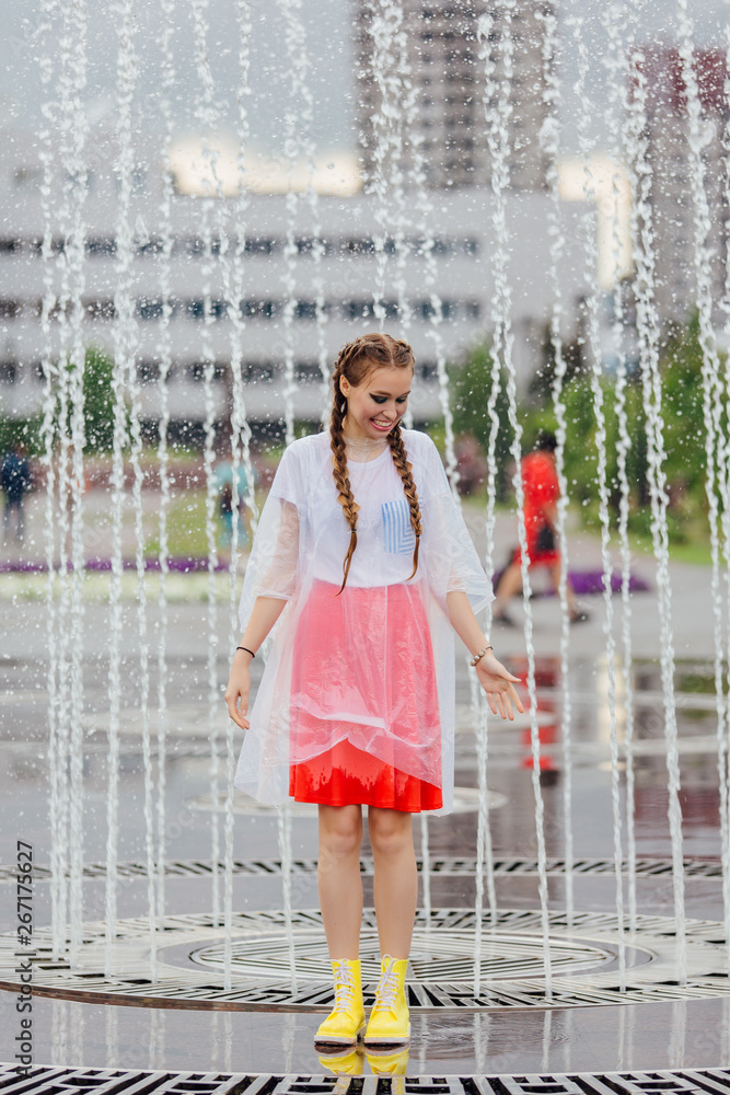 Wall mural Young wet pretty girl with two braids in yellow boots and with transparent umbrella stands inside of fountain.