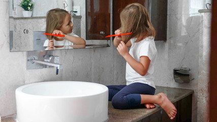 Small blonde girl is combing her hairs sitting on the table near the sink in front of the mirror in bathroom.