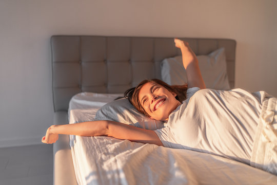 Happy girl waking up in the morning sunshine looking at sunrise sun in window excited to enjoy the day. Wake up energetic Asian woman lying in bed well rested from a good night sleep.