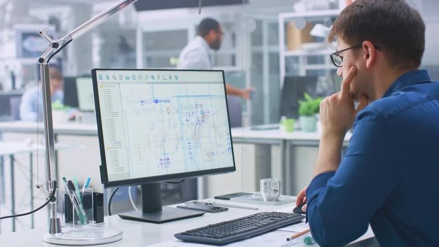 Over the Shoulder Shot of Engineer Working with CAD Software on Desktop Computer, Screen Shows Technical Drafts and Drawings. In the Background Engineering Facility Specialising on Industrial Design