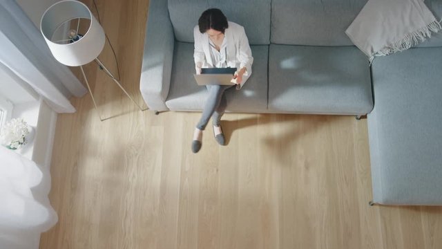 Young Woman Comes and Sits on a Couch, Working or Studying on a Laptop. Cozy Living Room with Modern Interior, Grey Sofa and Wooden Flooring. Top View Camera Footage.