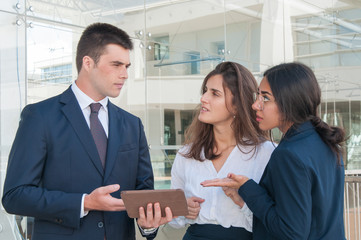 Serious young man in dark suite presenting colleagues data on tablet, they doubting idea, looking uncertain. Work, communication concept