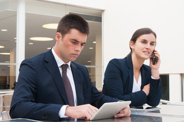 Business people using tablet and calling on phone at desk outdoors. Business man and woman wearing formal clothes and sitting at cafe table. Business and communication concept.