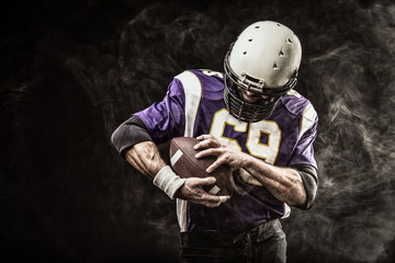 American football player holding ball in his hands in smoke. Black background, copy space. The...