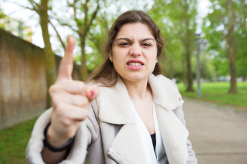 Angry nervous girl threatening at camera. Young woman in casual jacket standing in park and waging...