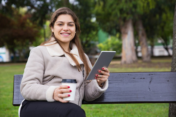Joyful Latin college girl with tablet drinking takeaway coffee in park. Young woman in warm jacket sitting on bench in park, holding gadget and smiling at camera. Internet connection outdoors concept