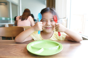 Obraz na płótnie Canvas Asian children cute hungry or kid girl holding colorful spoon and fork with dish for wait eat delicious food with smiling and happy for enjoy lunch or breakfast in morning at restaurant or food court