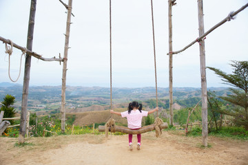 Child or back of kid girl playing wooden swing and happy fun or cheerful enjoy on nature holiday relax with landscape view in playground meadow field or amusement public park on summer travel vacation