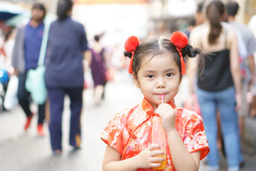 Asian child cute or Chinese kid girl stand and drinking orange juice by tube or straw in bottle for fresh and vitamin C with thirsty on hot weather summer in city street with wear chinese red dress