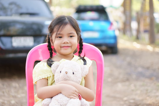 Asian child cute or kid girl pigtail smiling and hug teddy bear doll with happy to lovely toy on street or city park with car and sit on pink chair