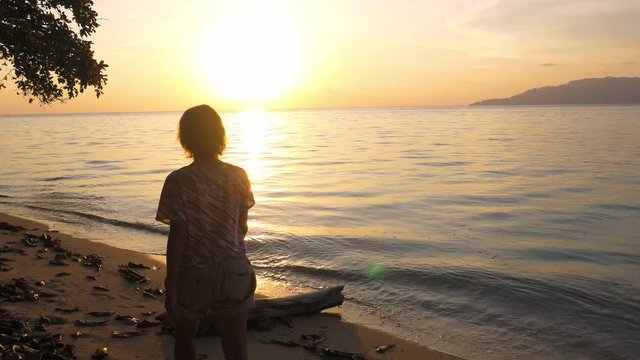 Woman relaxing on sand beach romantic sky at sunset, rear view, golden cloudscape, real people. Pulau Hatta Banda Islands Moluccas Maluku Indonesia