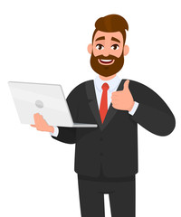 Happy young bearded business man holding/showing a latest new laptop and gesturing/making thumbs up sign with hand fingers. Like, good, positive, modern technology, digital device/gadget concept.