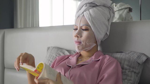 Attractive young woman with facial mask reading a book while sitting on the bedroom at home. Shot in 4k resolution