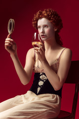 Medieval redhead young woman as a duchess in black corset and night clothes sitting on red background with a mirror and a glass of wine. Concept of comparison of eras, modernity and renaissance.