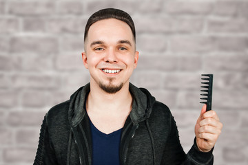 Young bearded man dressed in hoodie holding comb beside copy space on brick wall background