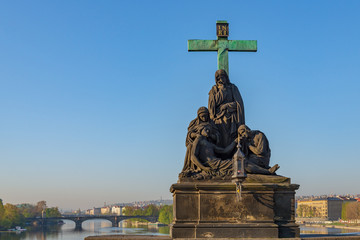 Fototapeta na wymiar Outdoor sunny view of Statuary of Pieta or Statue of the Lamentation of Christ, stand on balustrades of Charles Bridge, and background of Vltava river and Legions Bridge in Prague, Czech Republic.