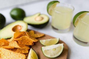Fresh avocado, lime, drink and nacho chips lying on marble background. Recipe for Cinco de Mayo party
