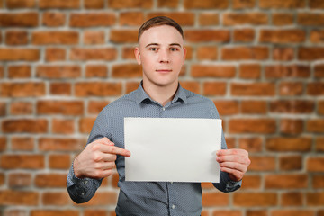 Portrait of young smiling caucasian guy showing blank with copy space on brick wall background