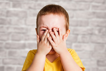 Crying baby boy in a yellow T shirt covers his face with hands and shouts, studio on brick wall background