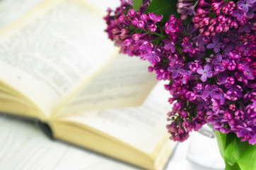 Naklejka na ściany i meble Lilac and a book. A bouquet of spring flowers against the background of an open book lying on a white wooden surface.