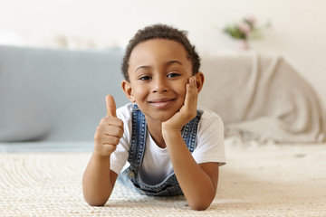 Picture of positive friendly looking eight year old Afro American boy lying on floor at home,...
